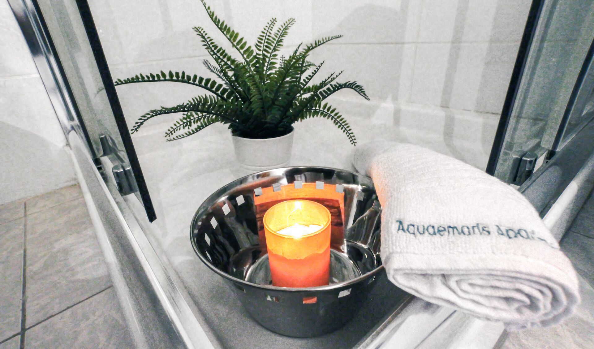 Um banheiro com box de vidro, samambaia em vaso, vela acesa em bandeja de metal e toalha com a inscrição "Aquamarine Spa".
