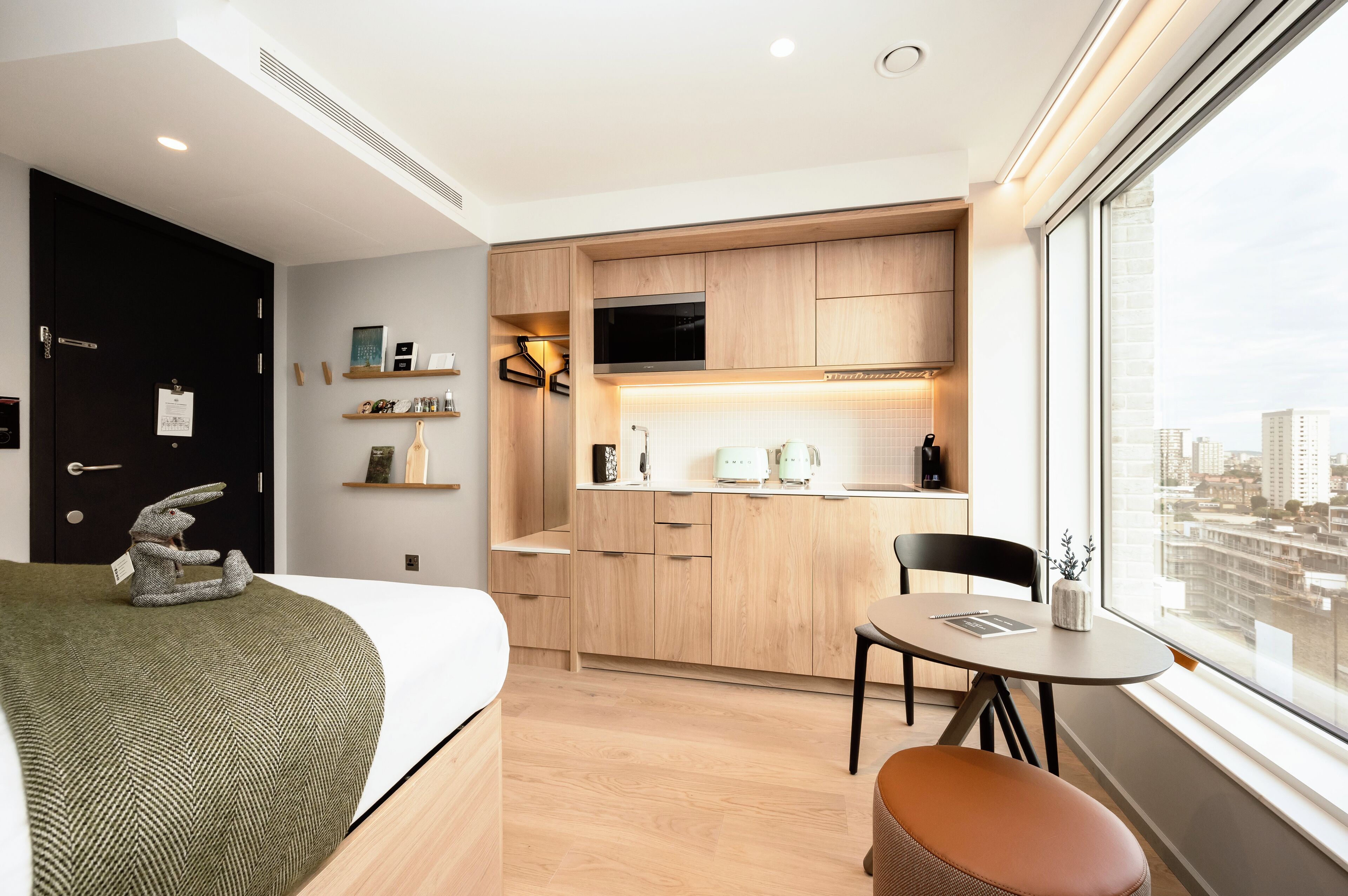 A modern hotel room with a bed, a small dining table, a chair, a kitchenette, and a large window offering a city view.