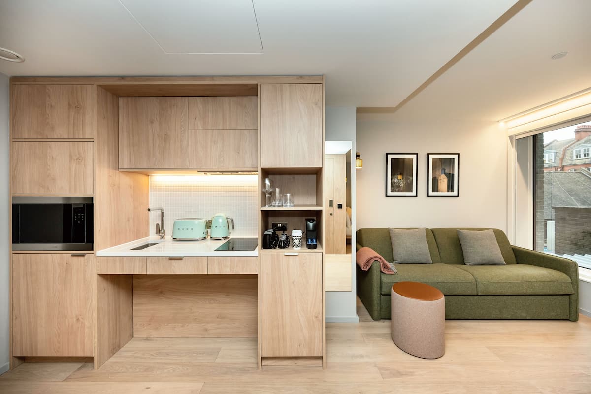 Une cuisine moderne avec des meubles en bois, un four à micro-ondes encastré et un plan de travail sur lequel se trouvent une bouilloire et une machine à café. On y trouve également un canapé vert et une fenêtre donnant sur des immeubles.