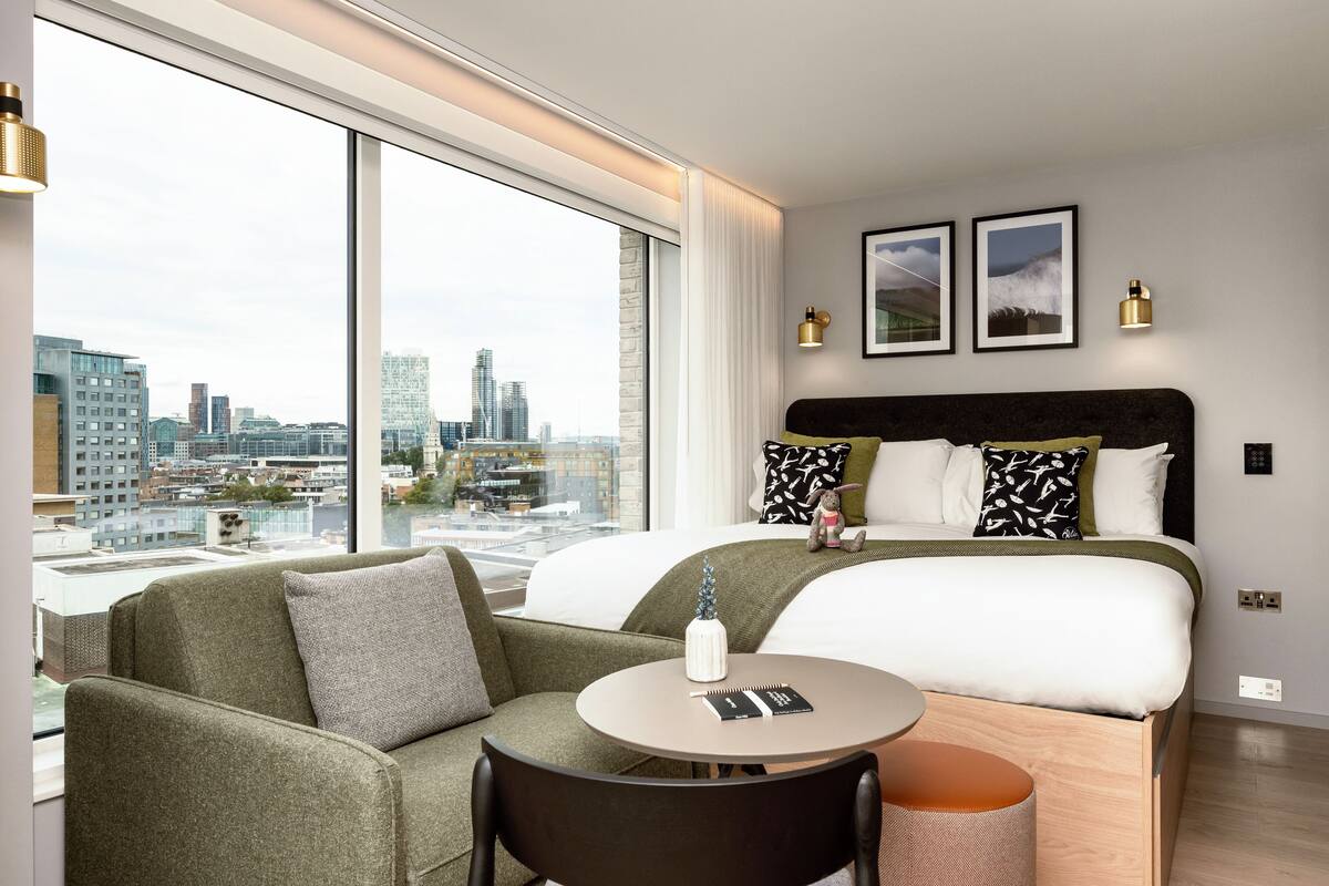 Une chambre d’hôtel moderne dotée d’un grand lit, d’un canapé, d’une table ronde et offrant une vue sur le paysage urbain grâce à de grandes fenêtres.