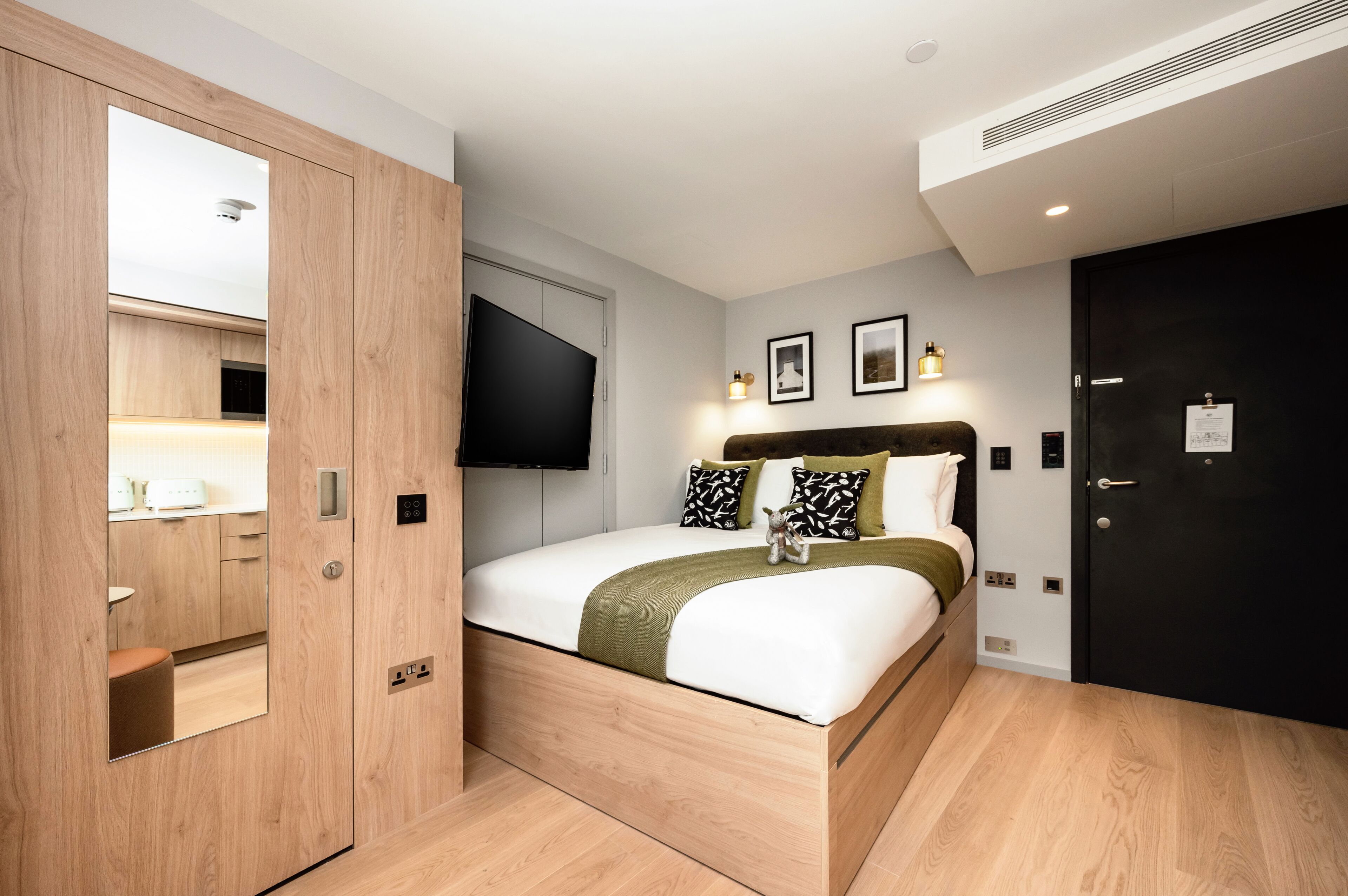 A modern bedroom with a wooden bed, a black wardrobe, a flat-screen TV, and a small table with a lamp.