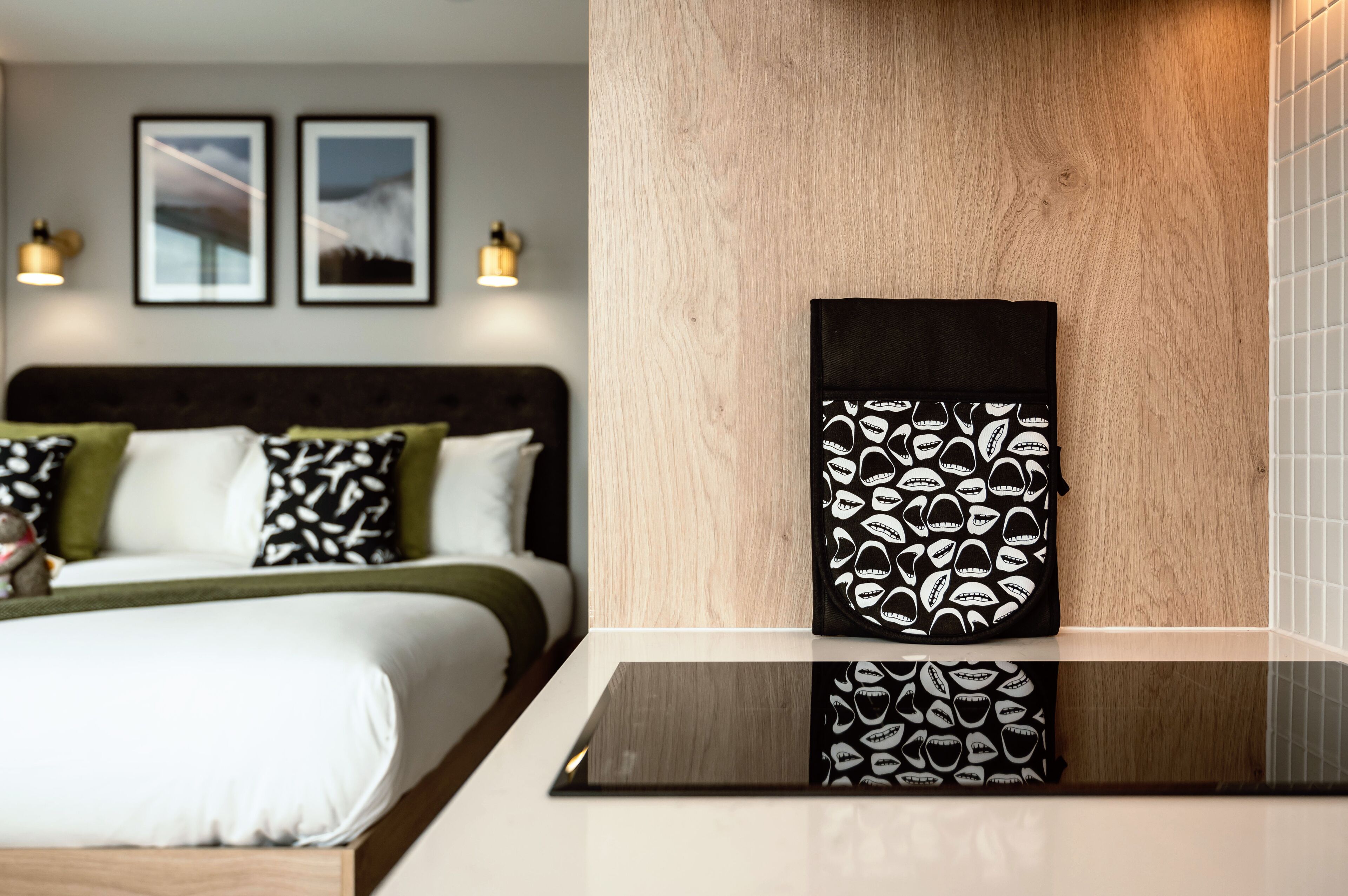 A modern hotel room with a wooden headboard, a bed with white and green bedding, and a black and white patterned pillow.