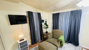 Una sala acogedora con sofá, reposapiés, televisor de pantalla plana, lámpara y cortinas.