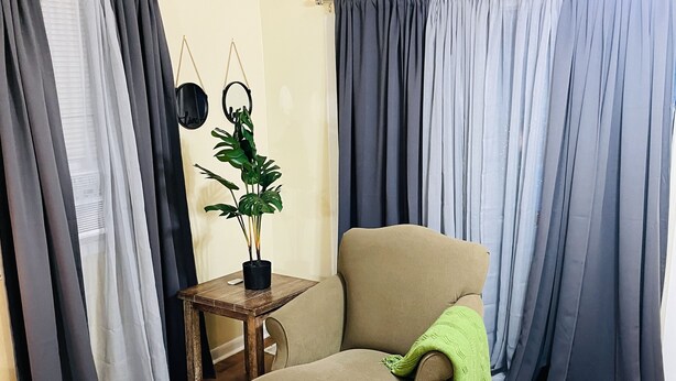 Una habitación con un sillón color beige, una mesita de madera con una planta en maceta y una cortina gris.