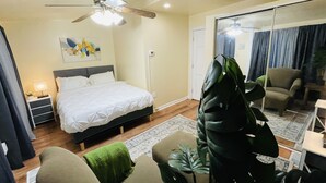 Un dormitorio con cama, ventilador de techo, mesita de noche, lámpara, espejo grande y una planta en maceta.