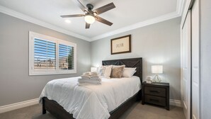 Quarto com cama, mesa de cabeceira, ventilador de teto, janela com persianas e uma frase inspiradora emoldurada na parede.