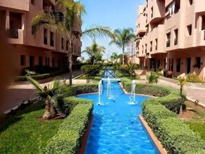 A courtyard with a central water feature, surrounded by well-maintained greenery and buildings with balconies. - Perfect apartment with a garden and pool—ideal for families with children (Marrakesch)