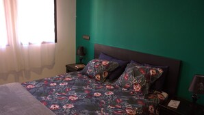 A bedroom with a floral bedspread, a headboard, a nightstand with a lamp, and a window with curtains.