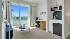 A modern living room with a sofa, a small round table, a kitchen area with a microwave, and a sliding glass door leading to a balcony with a city view. - Ocean & Bay Junior Suite in the Fontainebleau (Miami Beach)