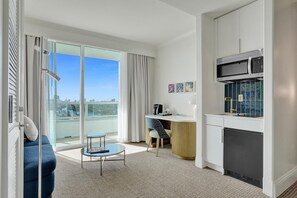 A modern living room with a sofa, a small round table, a kitchen area with a microwave, and a sliding glass door leading to a balcony with a city view.