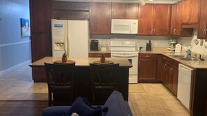 Cucina compatta con mobili in legno, un frigorifero bianco e un piano cottura. C'è una zona pranzo con due sedie e un tavolino, e un soggiorno con un divano e un tavolino da caffè.