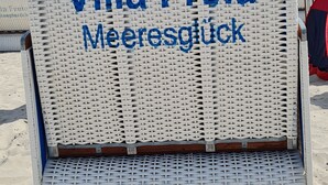 En flätad stol med texten ”Villa Freia Meeresblick” på, en mås som sitter ovanpå den, och en strand i bakgrunden.