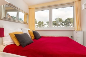 A bedroom with a red bedspread, a large window with yellow curtains, and a white bedside table. - Sea view in the 1st row Villa Freia Apartment Meeresglück with beach chair (Binz)