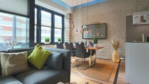A modern living room with a gray sofa, a dining area with a wooden table and chairs, and a kitchen with a refrigerator and shelves. - Luxury Loft Sylt - your special vacation home (Westerland)