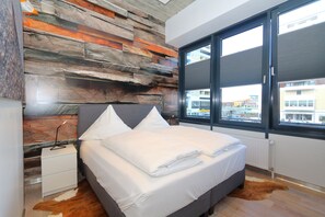 Un dormitorio moderno con una cama gris, ropa de cama blanca, una pared de acento de madera, una alfombra de piel de vaca y vistas de la ciudad a través de amplias ventanas.