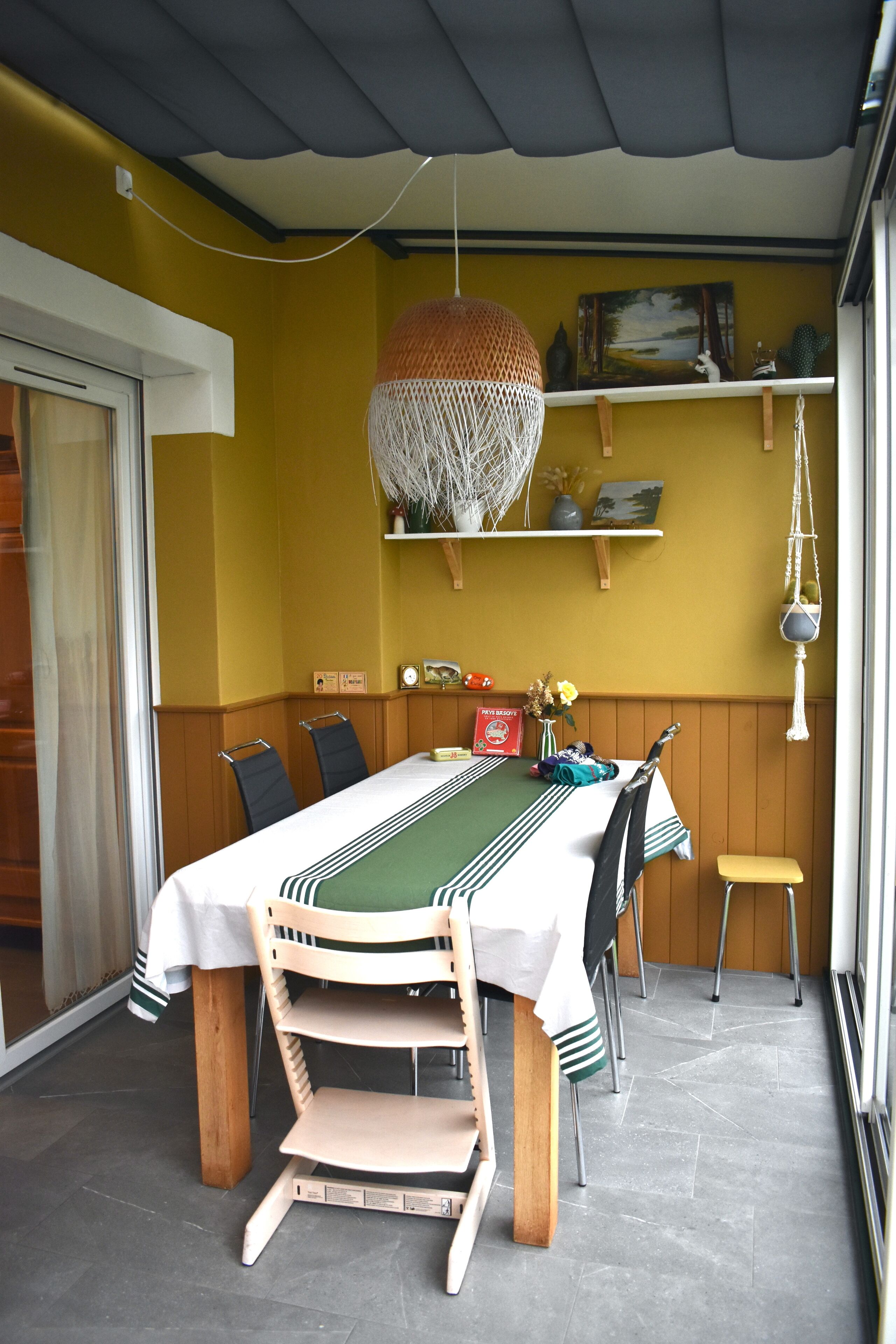 Een eetruimte met een tafel voor vier personen, houten stoelen, een hanglamp en een plank met decoratieve objecten.
