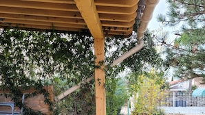 Uma área de estar coberta ao ar livre, com pérgola de madeira, uma cadeira de metal e um vaso com planta.