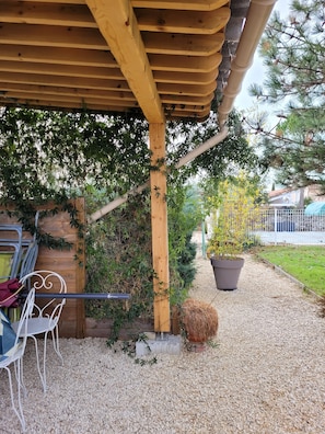 Un espace extérieur abrité, avec une pergola en bois, une chaise en métal et un pot de fleurs.