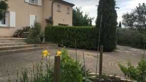 Um edifício residencial com telhado em telhas, entrada pavimentada e um jardim bem cuidado com flores e arbustos.