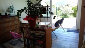 Quarto com estante de madeira cheia de jogos de tabuleiro, uma planta em vaso, uma cadeira e janela com vista para o verde lá fora.