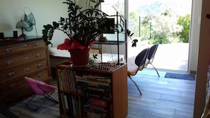 Une pièce avec une bibliothèque en bois remplie de jeux de société, une plante en pot, une chaise et une fenêtre donnant sur un paysage verdoyant.