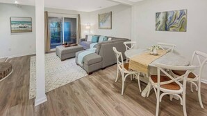 A modern living room with a sofa, dining table, and chairs, a large window, and artwork on the walls. - 5 Min to Beach, Renovated Townhome (Mount Pleasant)