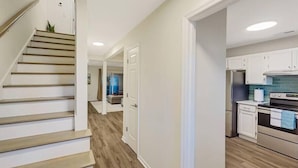 A modern kitchen with white cabinets and stainless steel appliances, a wooden staircase, and a hallway with a light-colored floor. - 5 Min to Beach, Renovated Townhome (Mount Pleasant)