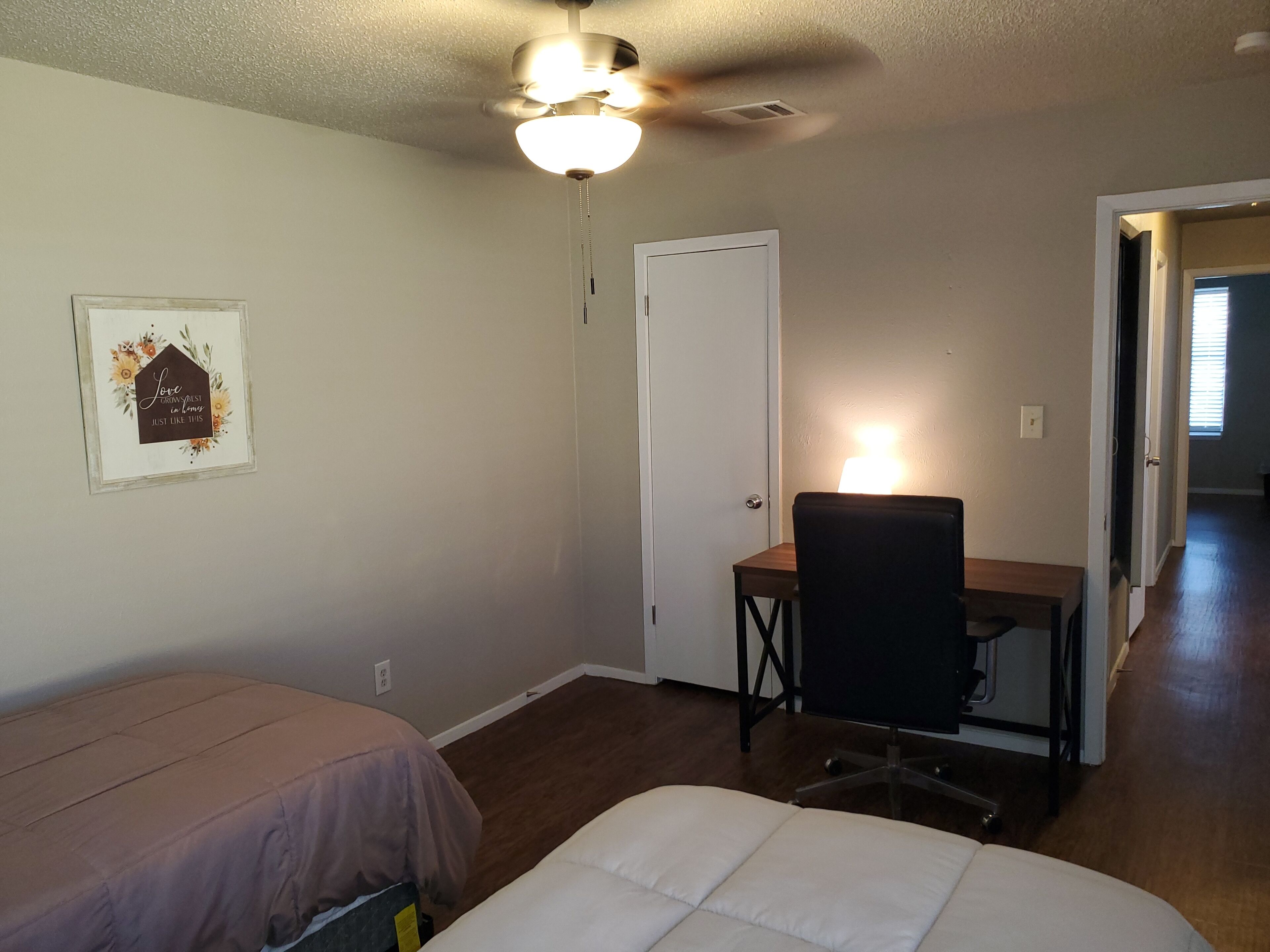 A bedroom with a bed, a desk with a chair, a ceiling fan, and a door.