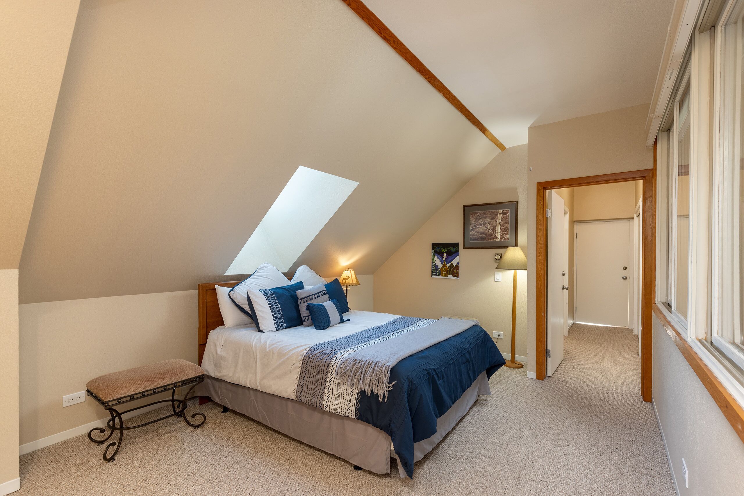 Een slaapkamer met een schuine kap, een bed met blauw beddengoed, een bankje en een lamp.