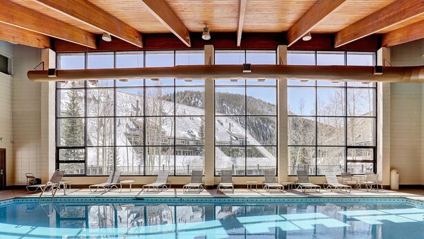 A large indoor swimming pool with multiple lounge chairs and a view of snowy mountains through large windows. - Beautiful Condo in the Pines, Mountain Views, Shared Pool & Hot Tub (Keystone)