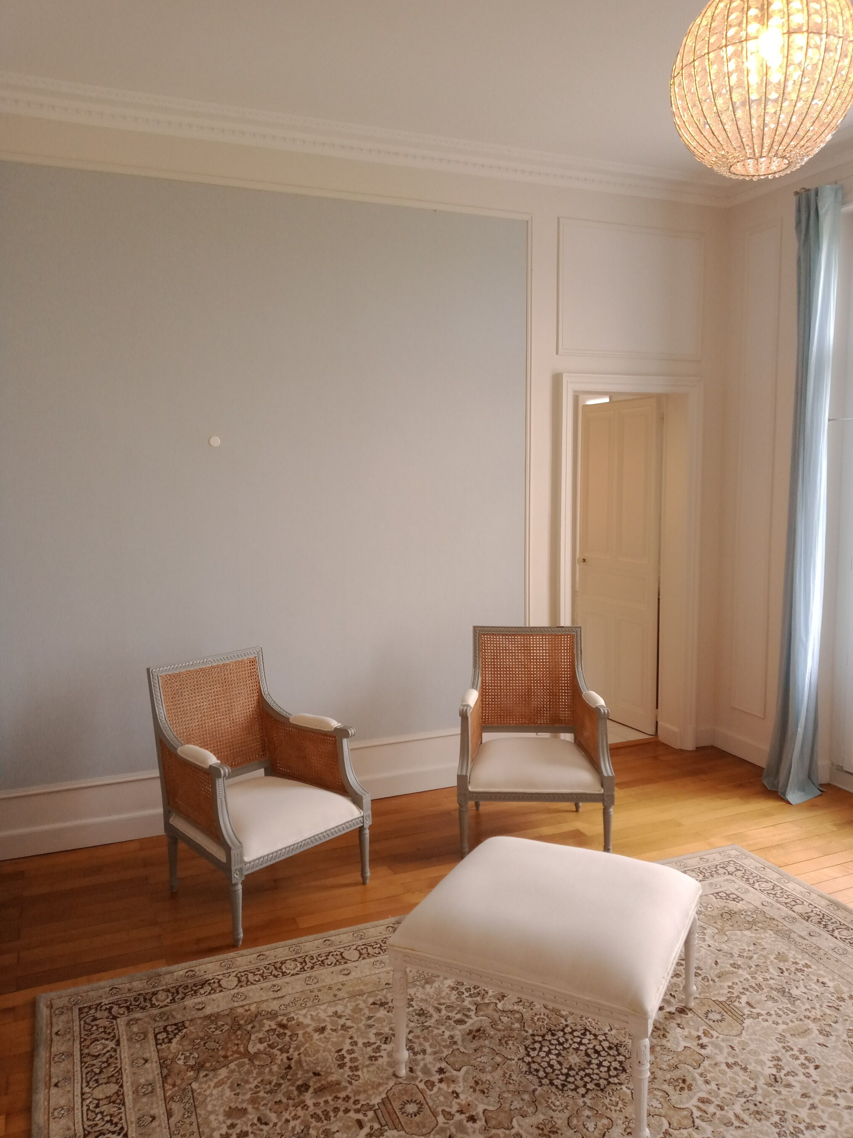 Una habitación con dos sillones de mimbre, un reposapiés blanco y una alfombra estampada sobre piso de madera.