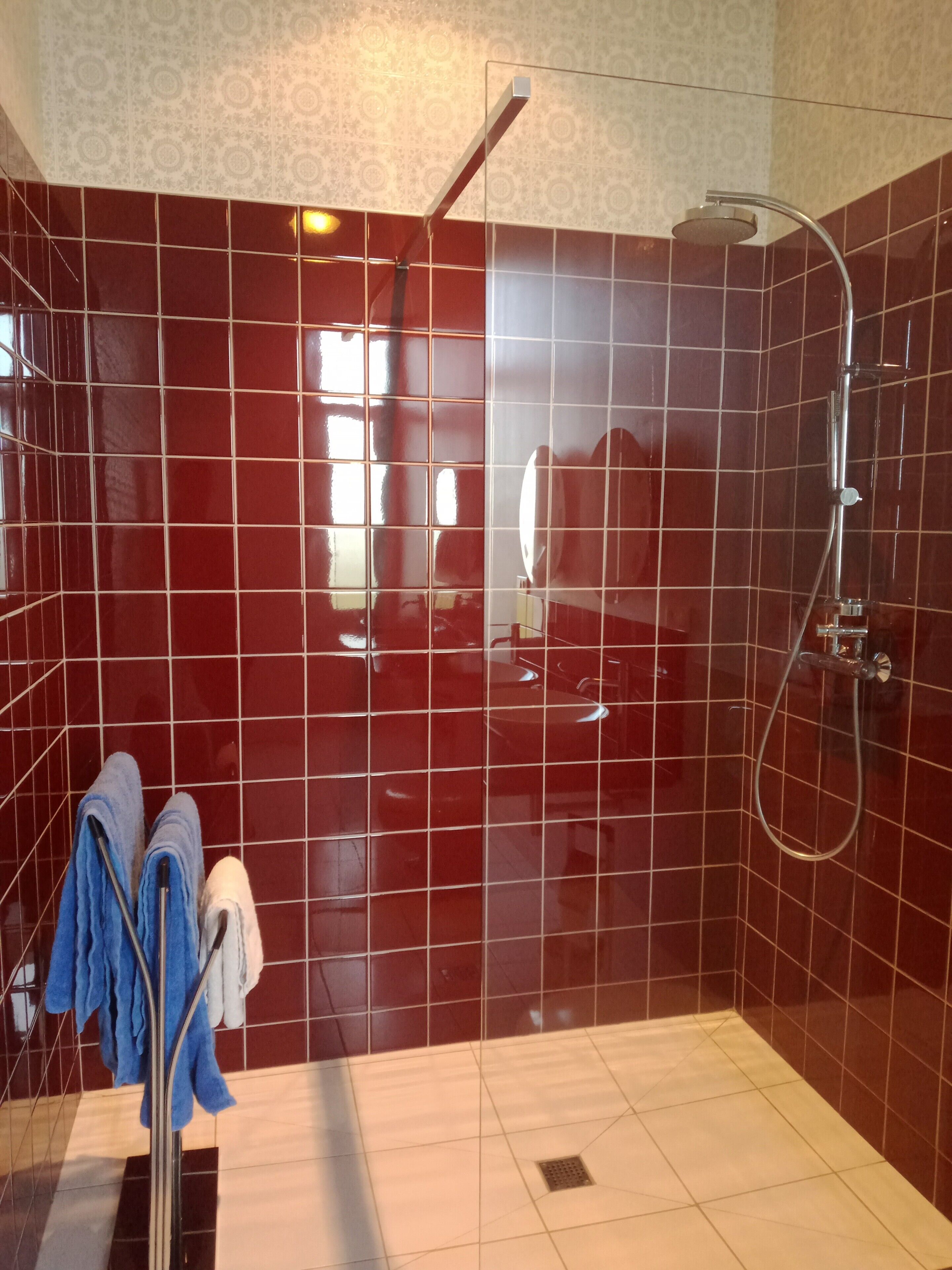 Ducha a pie con azulejos rojos, puerta de cristal y un perchero con toallas.