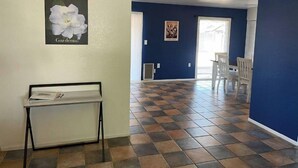 Une pièce avec un sol à carreaux, une table blanche recouverte de papiers, un tableau représentant une fleur, et un mur bleu.