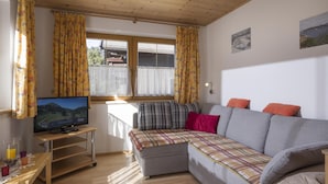 A cozy living room with a wooden ceiling, a sofa with a plaid blanket, a television on a stand, and a window with curtains. - Apartment 1 minute from the ski lift (Brixen im Thale)