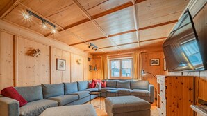 Ein gemütliches Blockhaushaus-Interieur mit Holzbalkendecke, einem großen grauen Sofa, einem kleinen grauen Hocker und einem Fernseher auf einem Holzschrank.