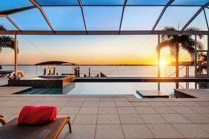 Ett område vid poolen med utsikt över solnedgången över vattnet, en möblerad sittplats under tak och en röd handduk på en stol.