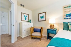 Une chambre à coucher comprenant un lit, une table de chevet, un fauteuil en osier, une commode et deux œuvres d’art encadrées accrochées au mur.
