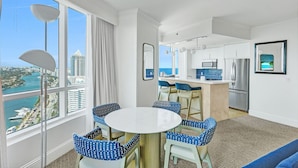 Une chambre d’hôtel moderne dotée d’une grande fenêtre donnant sur la ville, d’une cuisine équipée d’appareils électroménagers en acier inoxydable et d’un coin repas avec des chaises à motifs bleus.