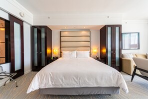 A large bed with a headboard, bedside tables with lamps, a chair, and a sofa in a modern hotel room. - Sorrento Ocean / Bay Jr Suite in the Fontainebleau (Miami Beach)