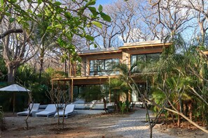 A modern house with a glass facade, surrounded by a garden with lounge chairs and palm trees.