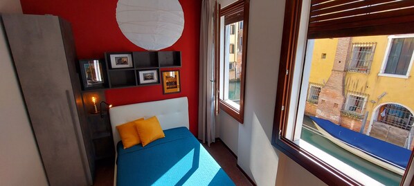 A bedroom with a bed, a red wall, a window with a view of buildings, and a white hanging light fixture.