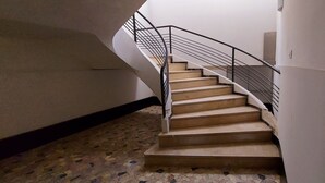 Un escalier en colimaçon avec une rampe en métal mène à l’étage supérieur. Les murs sont unis et le sol est recouvert de carreaux formant un motif.