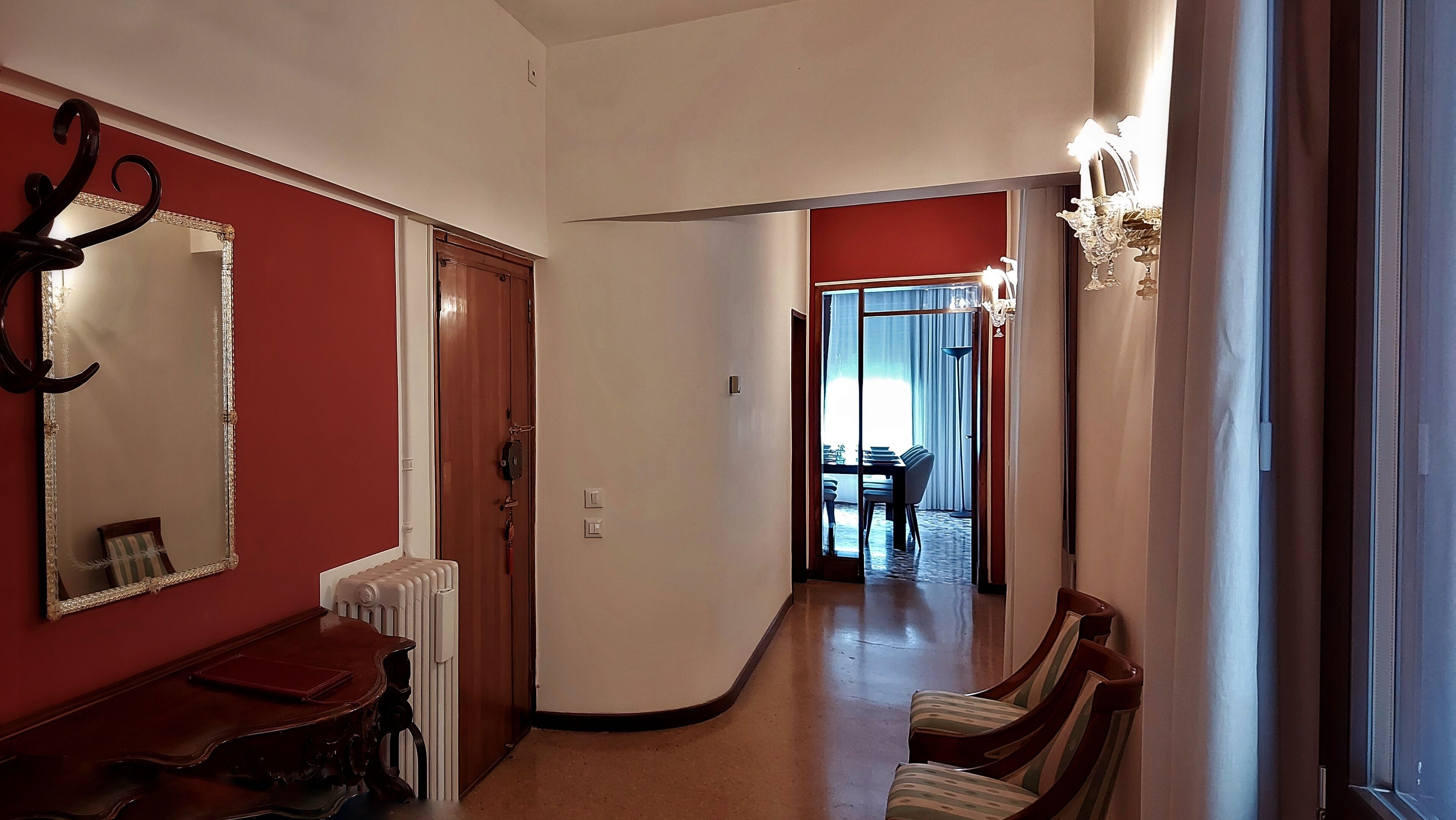 A hallway with a radiator, a wooden table, and a chair. There is a door with a mirror and a light fixture on the wall.