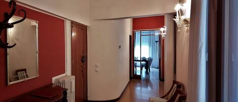 A hallway with a radiator, a wooden table, and a chair. There is a door with a mirror and a light fixture on the wall.