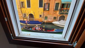Une vue à travers une fenêtre sur un canal, avec une gondole, des bâtiments et un auvent bleu.