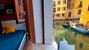 Une petite pièce comprenant un lit, une fenêtre et donnant sur un canal où naviguent des bateaux.