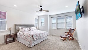 A bedroom with a bed, a nightstand, a chair, a ceiling fan, and a TV mounted on the wall.