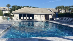 A swimming pool with a tiled deck, a covered area with chairs, and a clear blue sky.