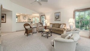A spacious living room with a beige sofa, two armchairs, a coffee table, and a ceiling fan. A large window provides natural light and a view of greenery outside.