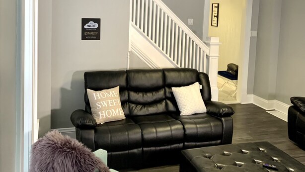 Ein modernes Wohnzimmer mit einem schwarzen Ledersofa, einem hellblauen Sessel und einem gepolsterten Hocker. Eine Treppe mit weißen Geländern führt zu einem Flur.